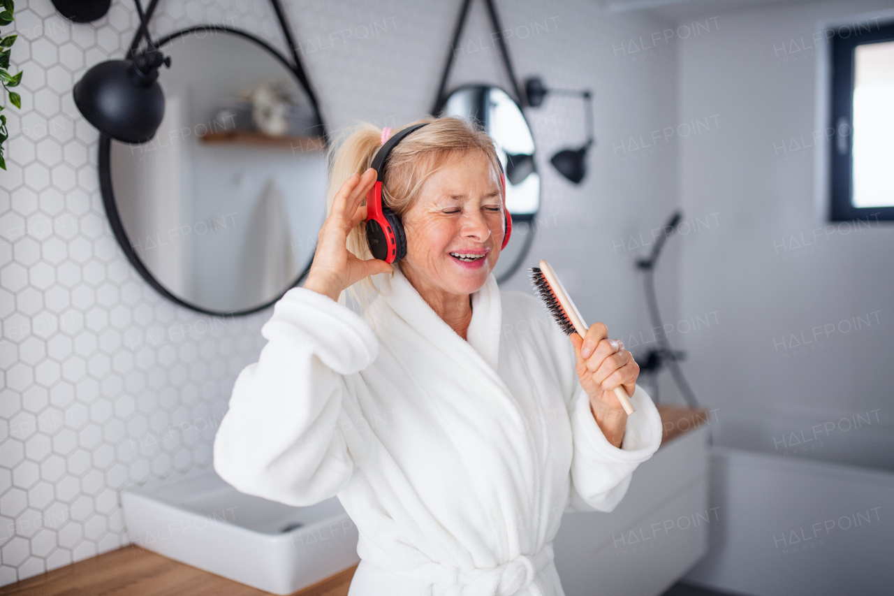 Portrait of senior woman with headphones and bathrobe indoors in bathroom at home, having fun.