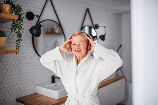 Portrait of senior woman with headphones and bathrobe indoors in bathroom at home.