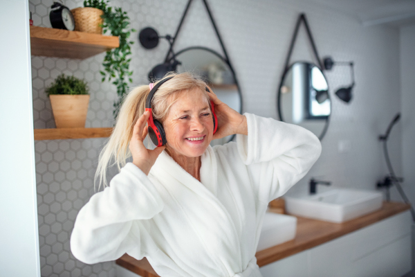 Portrait of senior woman with headphones and bathrobe indoors in bathroom at home.