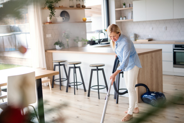 Portrait of happy senior woman with vacuum cleaner indoors at home, hoovering.