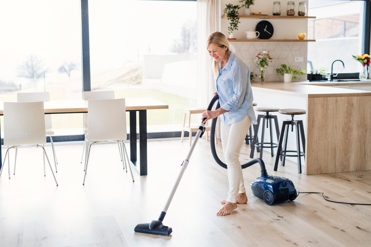 Portrait of happy senior woman with vacuum cleaner indoors at home, hoovering.