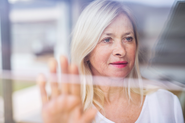 Portrait of depressed senior woman standing by window indoors at home. Shot through glass.