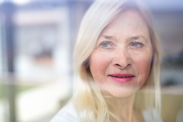 Close-up portrait of senior woman standing indoors at home. Shot through glass.