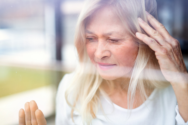 Portrait of depressed senior woman standing by window indoors at home. Shot through glass.