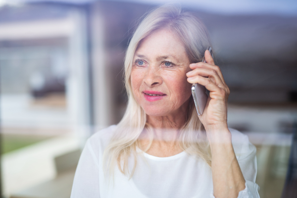 Portrait of senior woman standing indoors at home, using smartphone. Shot through glass.