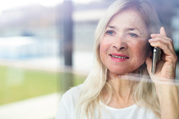 Portrait of senior woman standing indoors at home, using smartphone. Shot through glass.