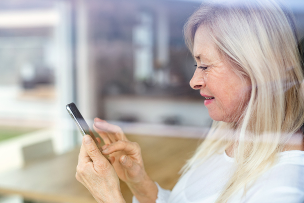 Portrait of senior woman standing indoors at home, using smartphone. Shot through glass.