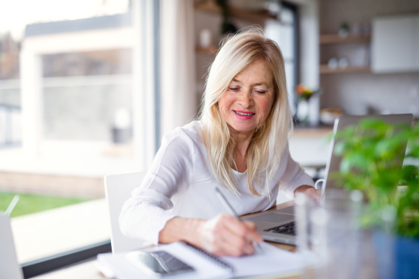 Senior woman with laptop indoors in home office, writing when working.