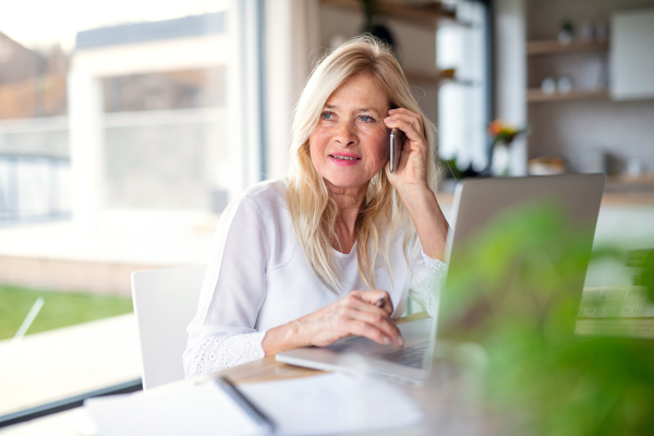 Portrait of senior woman with smartphone and laptop indoors in home office, working.