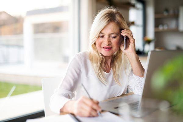 Portrait of senior woman with smartphone and laptop indoors in home office, working.