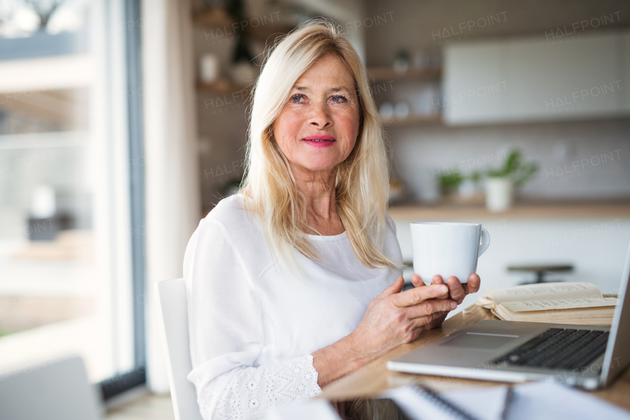 Portrait of senior woman with coffee and laptop indoors in home office, working.