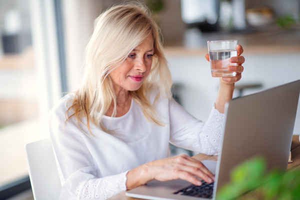 Portrait of senior woman with glass of water and laptop indoors in home office, working.