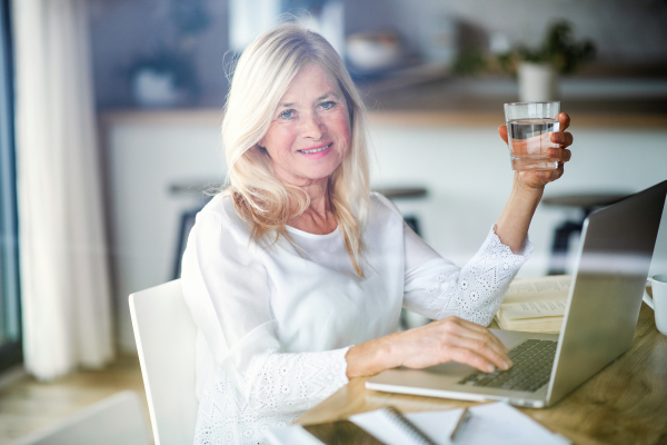 Senior woman with water and laptop indoors in home office, working. Shot through glass.