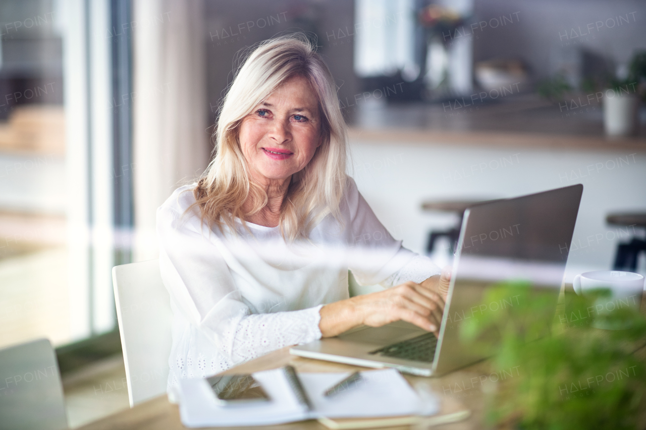 Senior woman with laptop indoors in home office, working. Shot through glass.