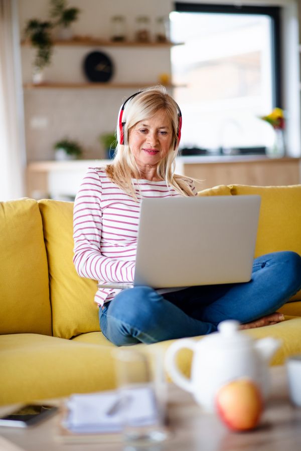 Senior woman with headphones and laptop sitting on sofa indoors at home, relaxing.