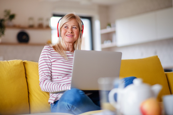 Senior woman with headphones and laptop sitting on sofa indoors at home, relaxing.