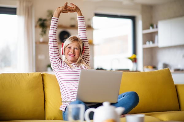 Senior woman with headphones and laptop sitting on sofa indoors at home, relaxing.