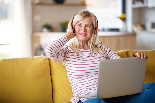 Senior woman with headphones and laptop sitting on sofa indoors at home, relaxing.