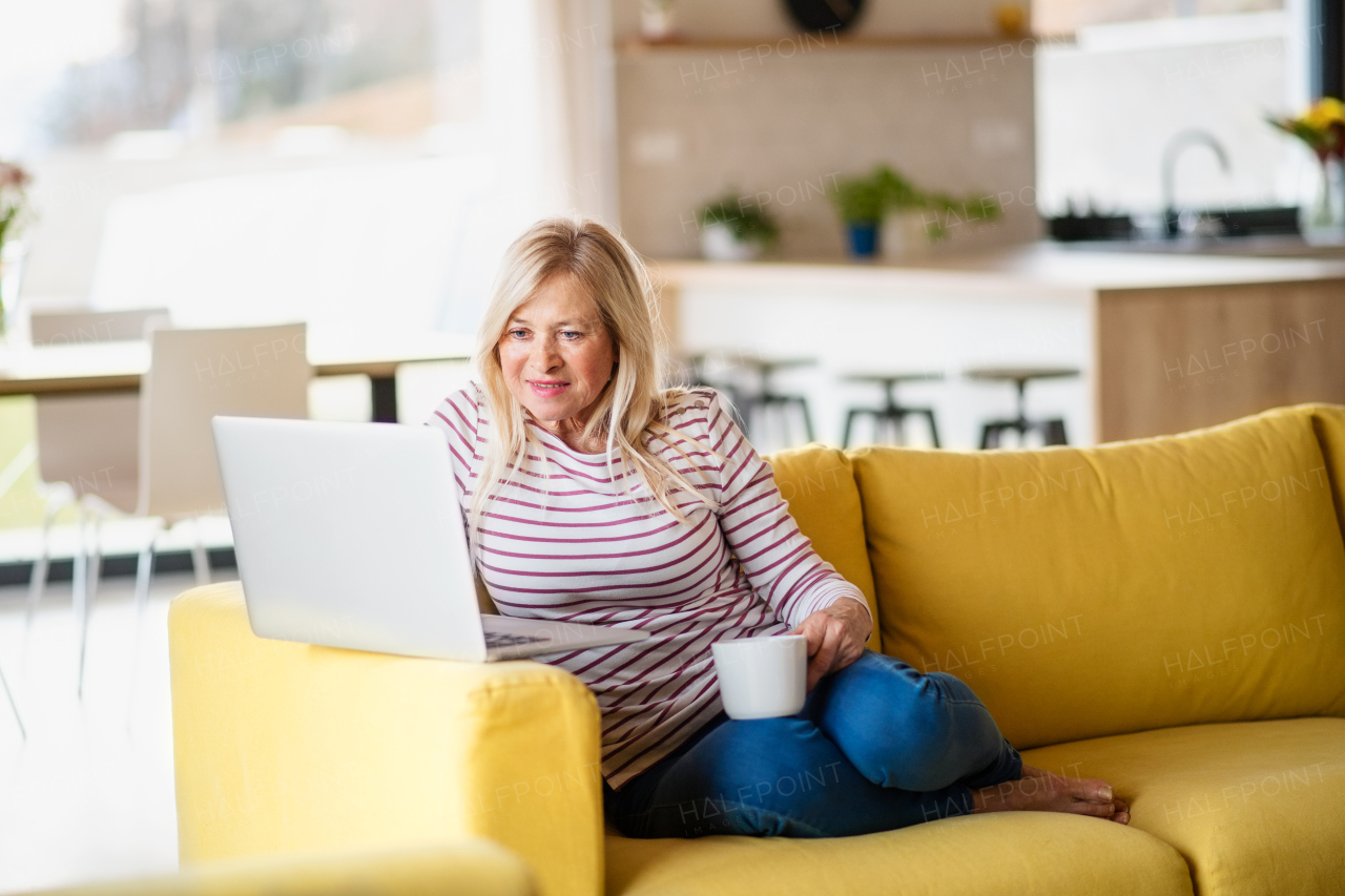 Senior woman with coffee and laptop indoors at home, relaxing on sofa.