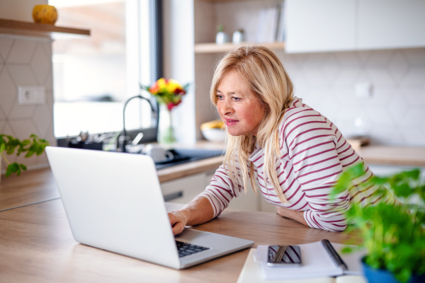 Front view of senior woman with laptop indoors in kitchen home office, working.