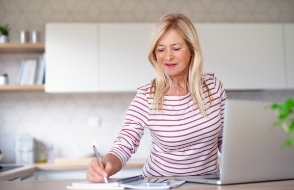 Front view of senior woman with laptop indoors in kitchen home office, working.