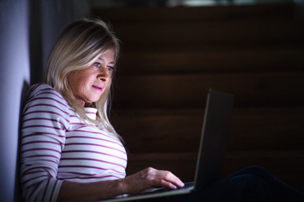 Senior woman with laptop indoors at home in the evening, working. Copy space.