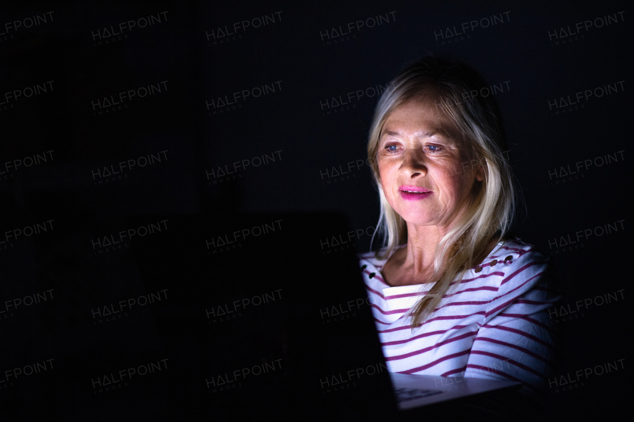 Senior woman with laptop indoors at home working late at night. Copy space.