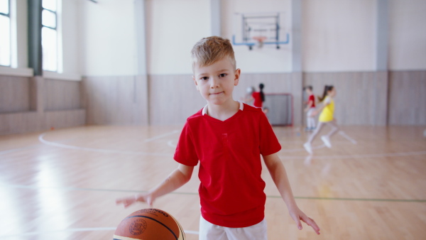 A cheerful schoolboy dribbling with basketball indoors at gym at school.