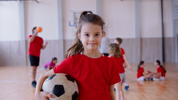 A small multiracial girl standing with ball indoors in gym class, physical education concept