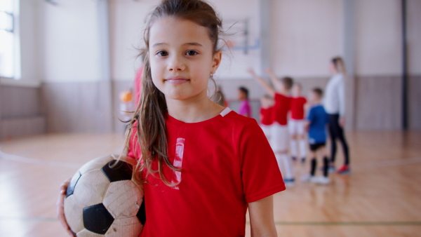 A small girl standing with ball indoors in gym class, physical education concept