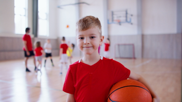 A cheerful schoolboy dribbling with basketball indoors at gym at school.