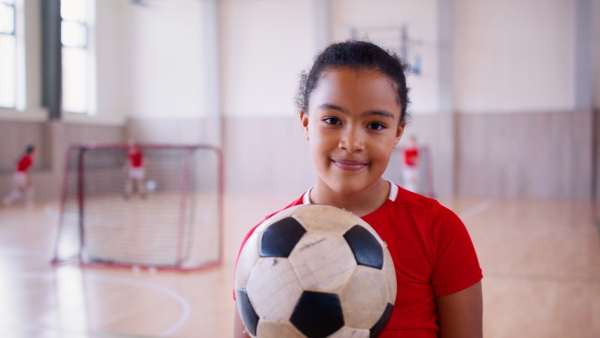 A small multiracial girl standing with ball indoors in gym class, physical education concept