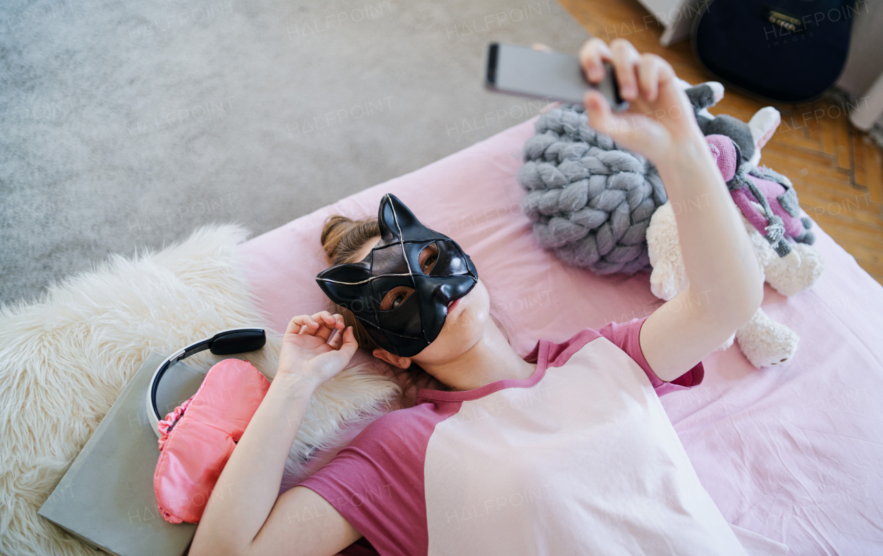 Top view of young girl with cat mask on bed, taking selfie. Online dating concept.