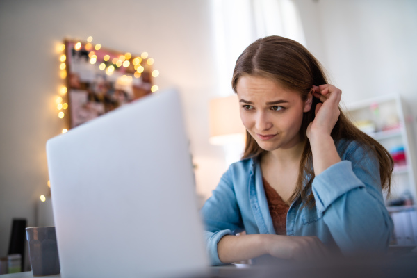 Sad and worried young girl with laptop and smartphone sitting on bed indoors, internet abuse concept.