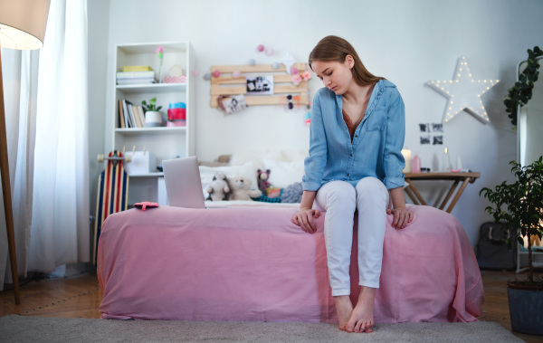 Sad and worried young girl with laptop sitting on bed indoors, internet abuse concept.