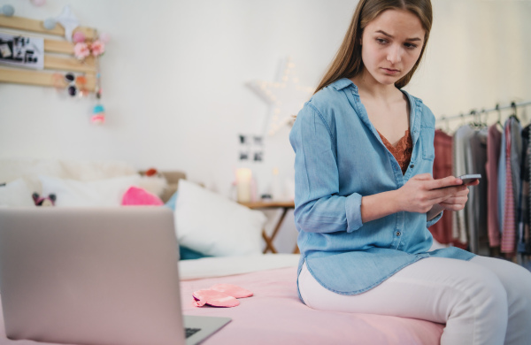 Sad and worried young girl with laptop and smartphone sitting on bed indoors, internet abuse concept.