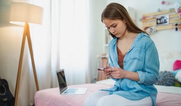 Sad and worried young girl with laptop and smartphone sitting on bed indoors, internet abuse concept.