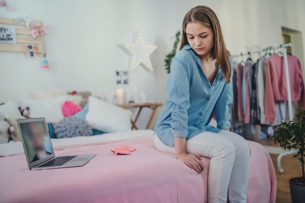 Sad and worried young girl with laptop sitting on bed indoors, internet abuse concept.