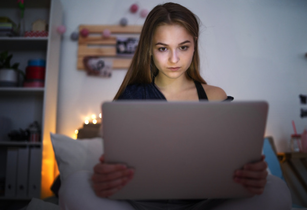 Sad and worried young girl with laptop and smartphone sitting on bed indoors, internet abuse concept.