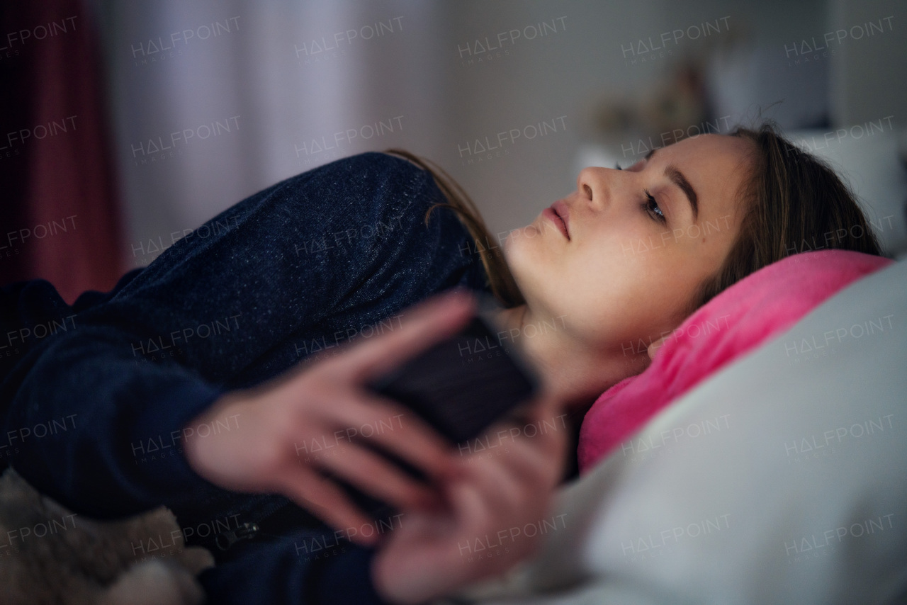 Sad young girl with smartphone lying indoors on bed, online dating and internet abuse concept.