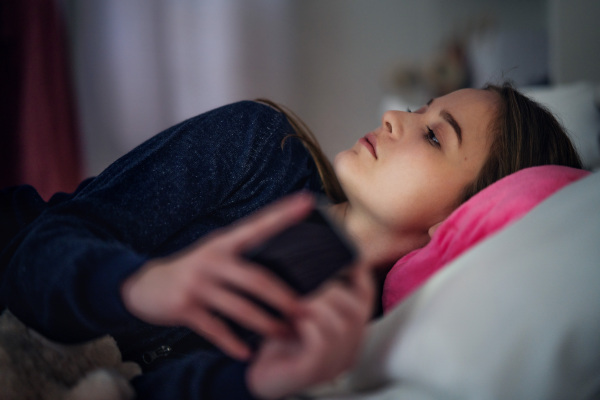 Sad young girl with smartphone lying indoors on bed, online dating and internet abuse concept.