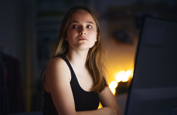 Sad and worried young girl with computer sitting indoors, internet abuse concept.