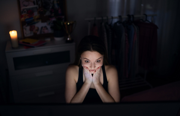 Front view of scared and worried young girl with laptop sitting indoors, internet abuse concept.