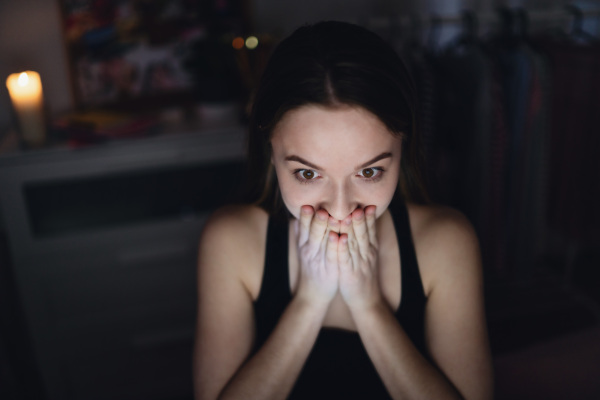 Front view of scared and worried young girl with laptop sitting indoors, internet abuse concept.