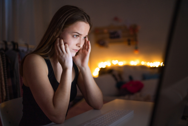 Sad and worried young girl with computer sitting indoors, internet abuse concept.