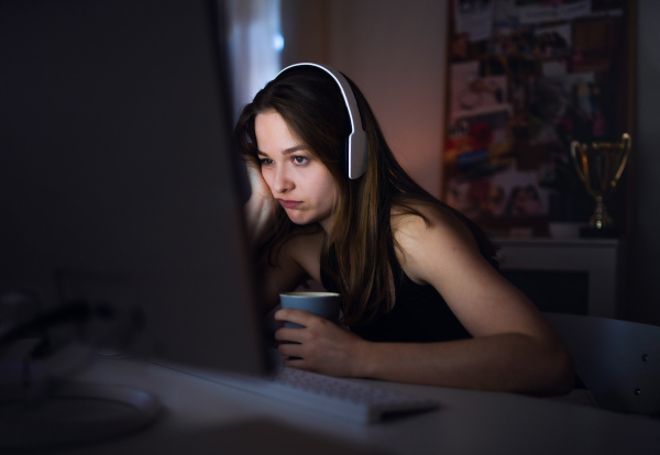 Side view of bored young girl with headphones and computer sitting indoors, online chatting concept.