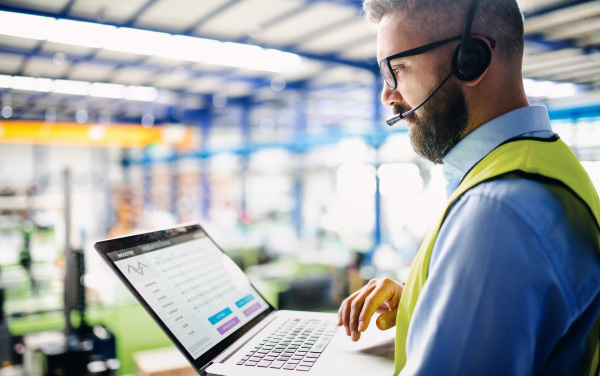 Side view of technician or engineer with headset and laptop standing in industrial factory, working.