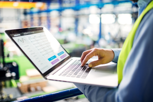 Unrecognizable technician or engineer with laptop standing in industrial factory, working.