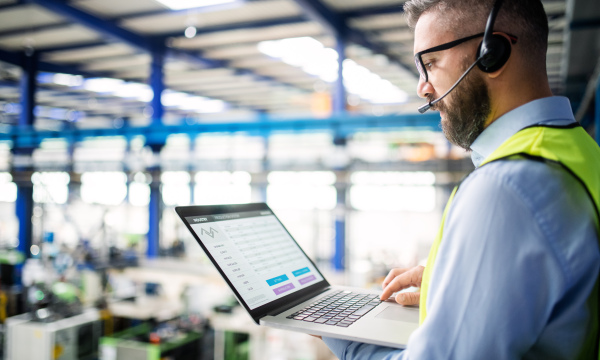 Side view of technician or engineer with headset and laptop standing in industrial factory, working.