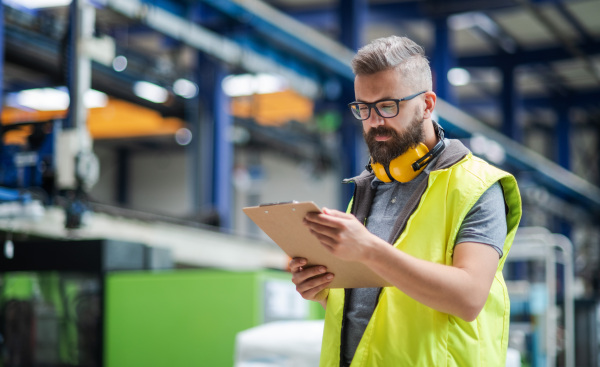 Technician or engineer with protective headphones standing in industrial factory, working.